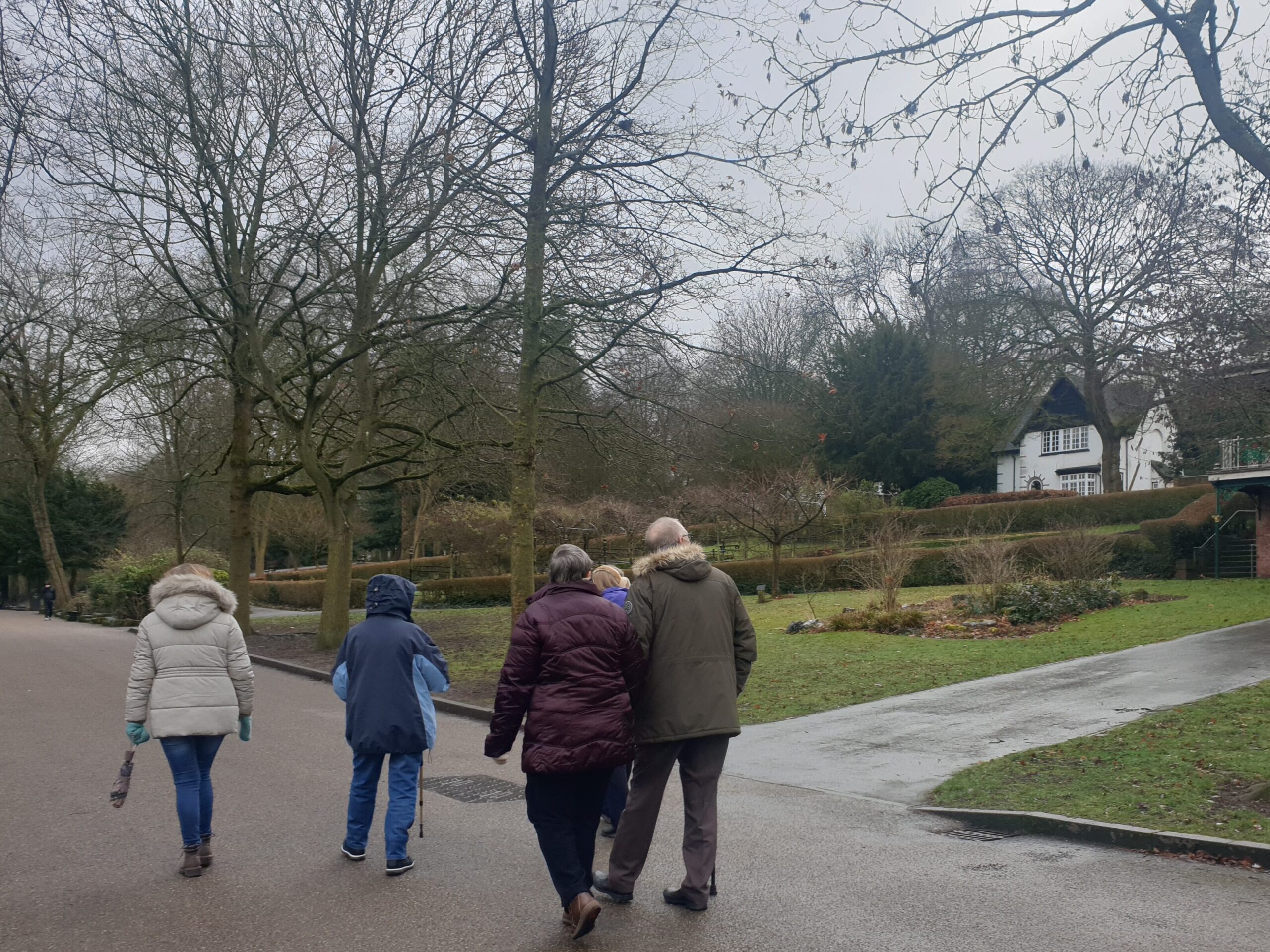A photo of 5 people wrapped up in winter coats taking a walk around Walsall Arboretum. The gardens are tended and the trees are without leaves. In the background, there is a historic building.