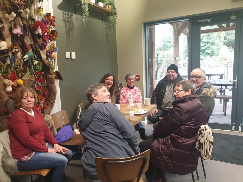 A mixed group of Carers are sat around a table in a coffee shop. There are coffees on the table and many of the Carers are still wearing their coats after the walk. On the wall behind them, there is a three dimensional textile art piece. This piece is a scene of the outdoors, with plants, flowers, insects and animals.