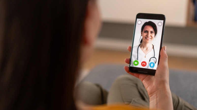 A person is sat comfortably while holding their phone. The phone screen shows that they are on o video call to their doctor/