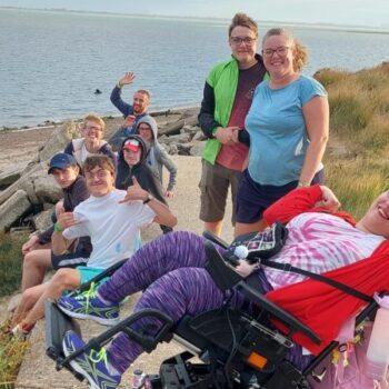 A mixed age group of people including several with various additional needs and carers photographed on a coastal path with a beach and the sea in the background. All are facing the camera with happy expressions, waving and joyful gestures.