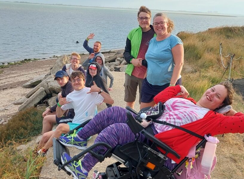 A mixed age group of people including several with various additional needs and carers photographed on a coastal path with a beach and the sea in the background. All are facing the camera with happy expressions, waving and joyful gestures.